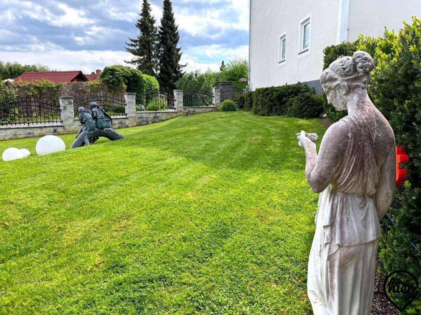 Gepflegter Garten mit weitläufiger Rasenfläche, einer klassischen Statue und einem Zaun im Hintergrund.