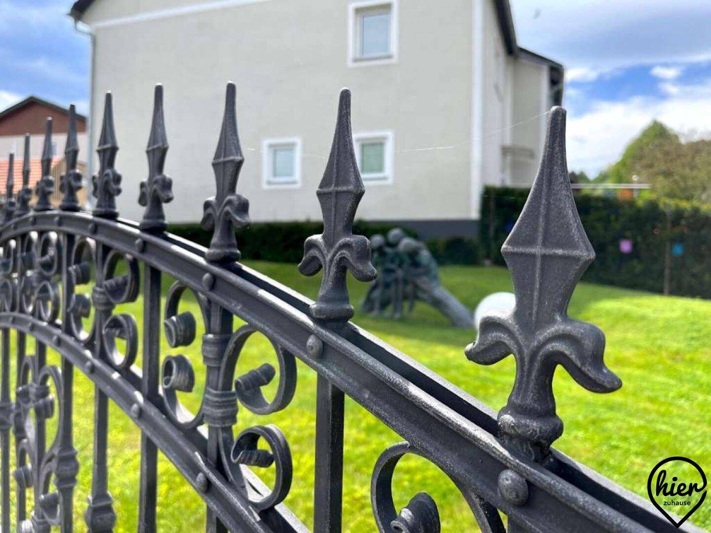 Detailaufnahme eines kunstvollen schmiedeeisernen Zauns mit einem Haus und Garten im unscharfen Hintergrund.