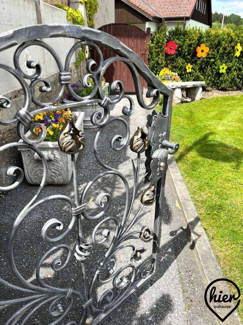 Ein kunstvoll geschmiedetes Gartentor öffnet sich zu einem gepflegten Gartenbereich mit Blumen.