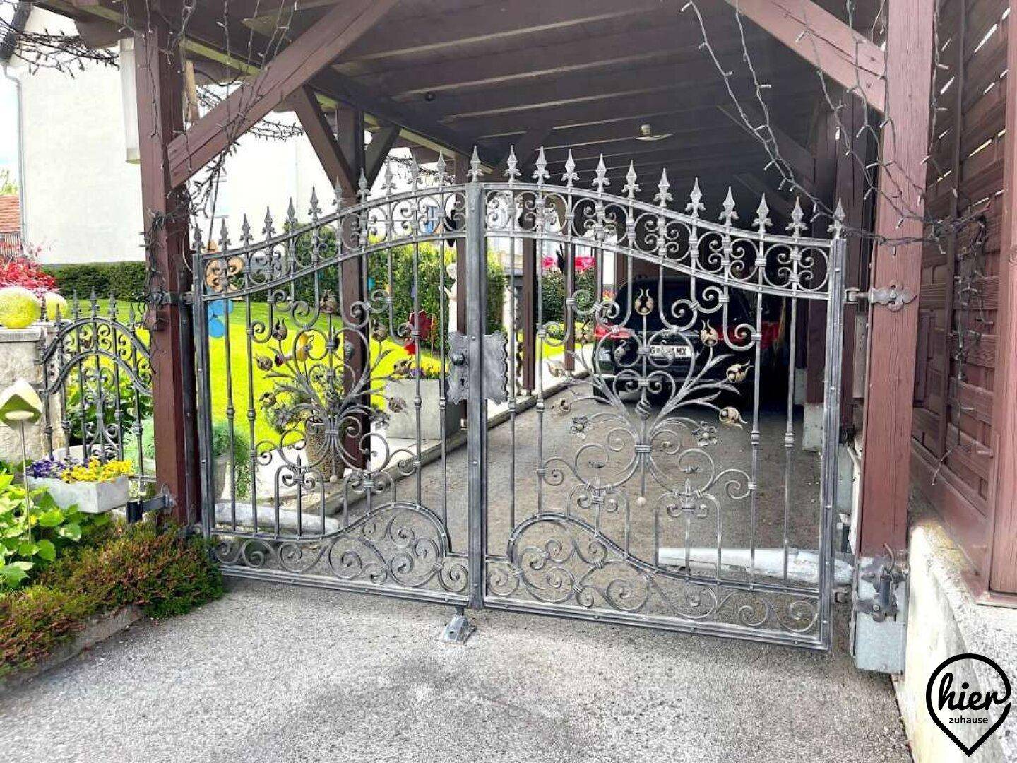 Ein großes, schmiedeeisernes Doppeltor führt zum überdachten Carport und dem Eingangsbereich des Hauses.