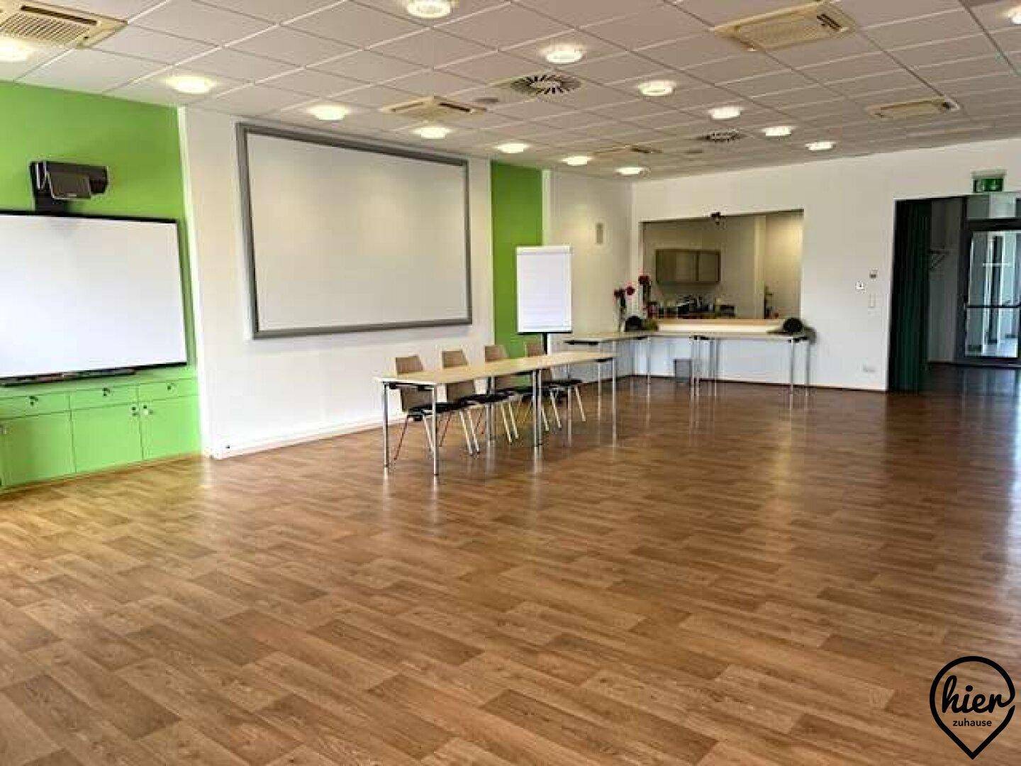 Großer Konferenzraum mit Holzboden, Projektionsflächen und einer Cateringküche im Hintergrund.