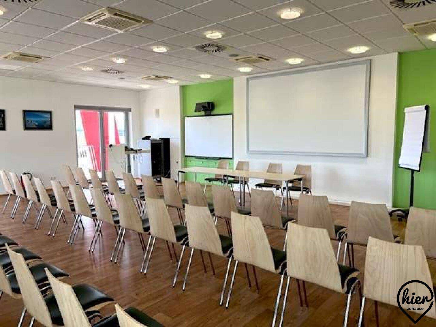 Konferenzraum mit Reihenbestuhlung, Holzboden und Blick auf die Projektionsflächen.