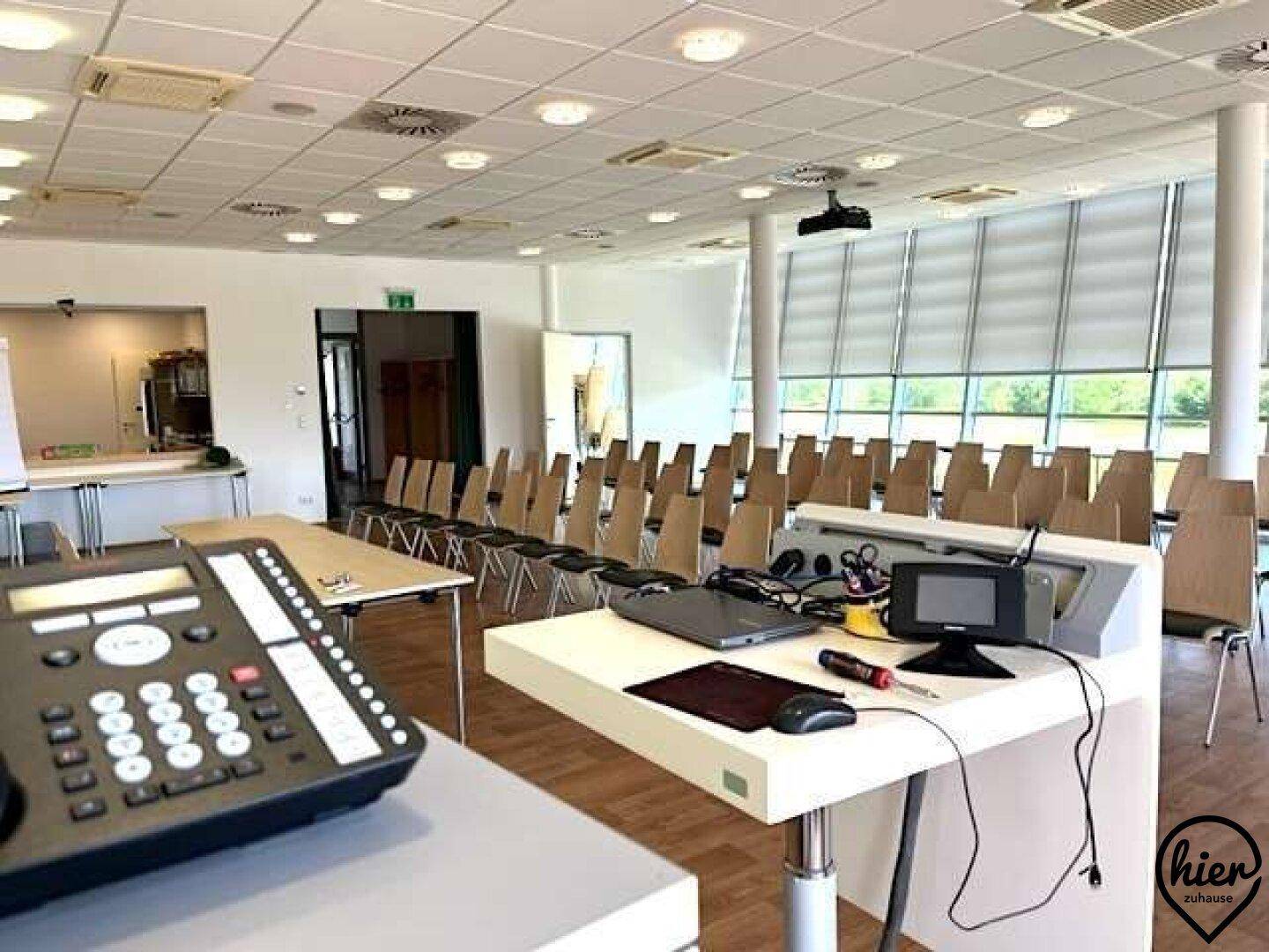 Konferenzraum mit Reihenbestuhlung, großen Fenstern und einem Rednerpult im Vordergrund.