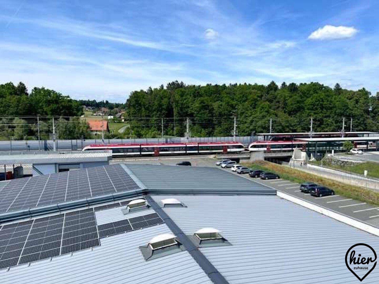 Blick auf eine Bahnstrecke mit Zügen, Parkplätzen und bewaldeter Umgebung.