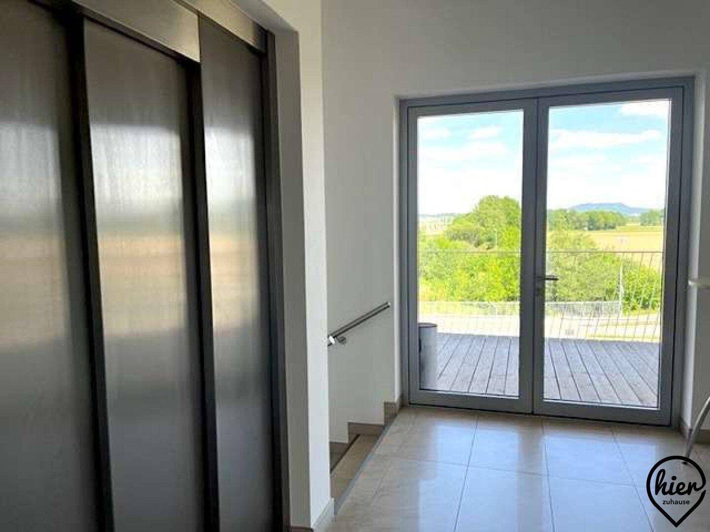 Heller Flur mit Fliesenboden, Aufzug und Zugang zu einer Außenterrasse mit Weitblick.