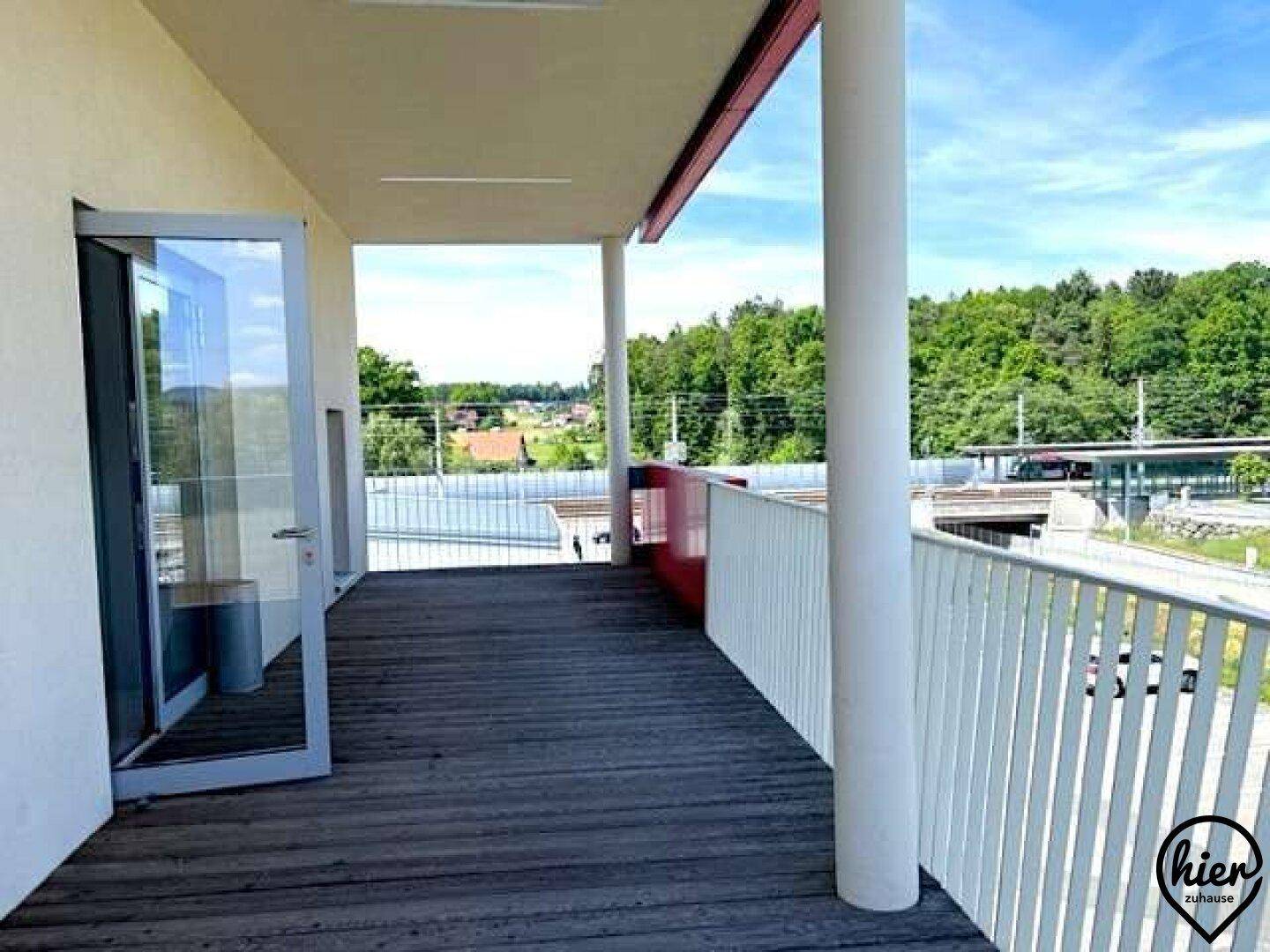 Langer Balkon mit Holzboden und Blick auf Bäume, eine Straße und weitere Gebäude in der Ferne.