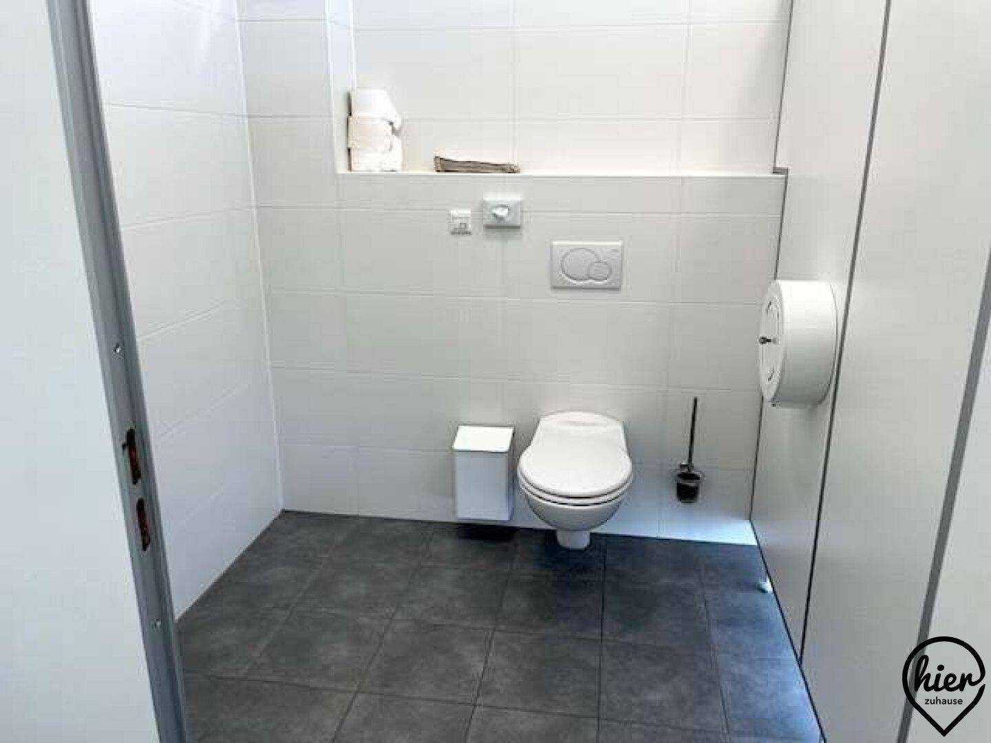 Toilettenkabine in einem öffentlichen Waschraum mit weißen Fliesenwänden und dunklem Fliesenboden.