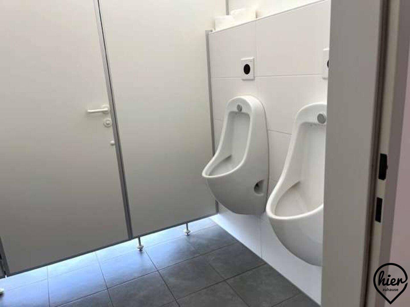 Öffentlicher Toilettenbereich mit zwei Urinalen, weißen Fliesenwänden und dunklem Fliesenboden.