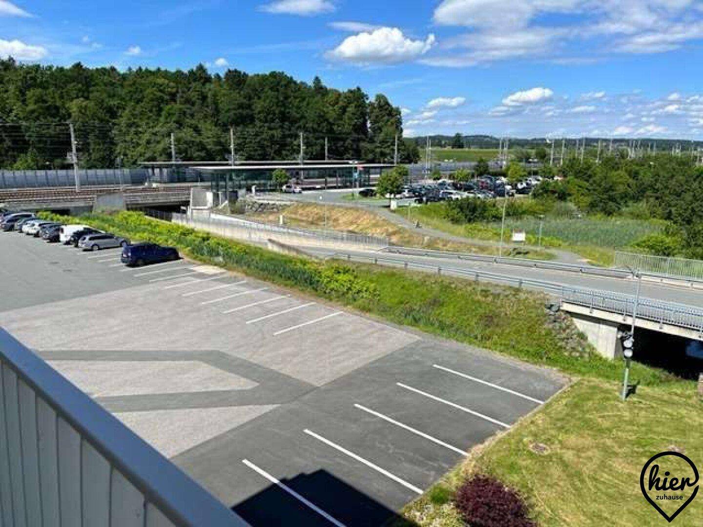 Großer Parkplatz mit markierten Stellplätzen, umgeben von Grünflächen, mit Straße und entfernten Gebäuden.