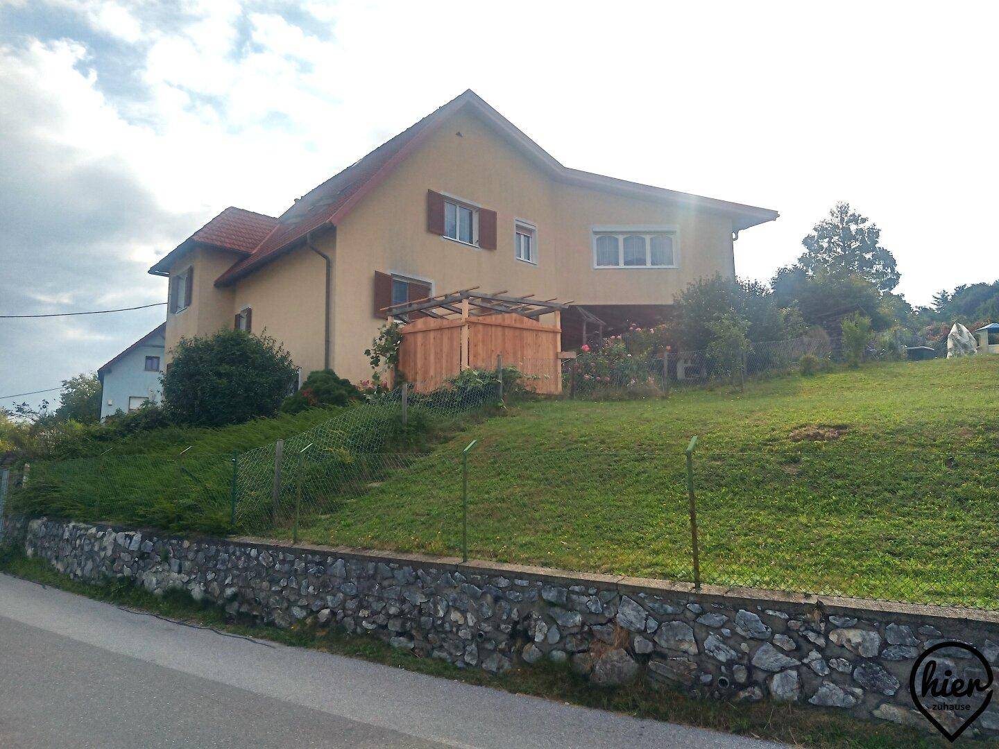 Einfamilienhaus mit gelber Fassade und großem, grünem Hanggrundstück unter bewölktem Himmel.