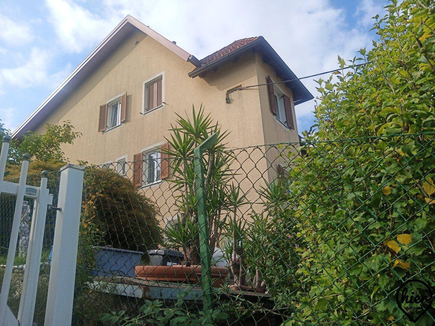Gelbes Haus mit braunen Fensterläden, umgeben von üppigem Grün und einem Maschendrahtzaun.