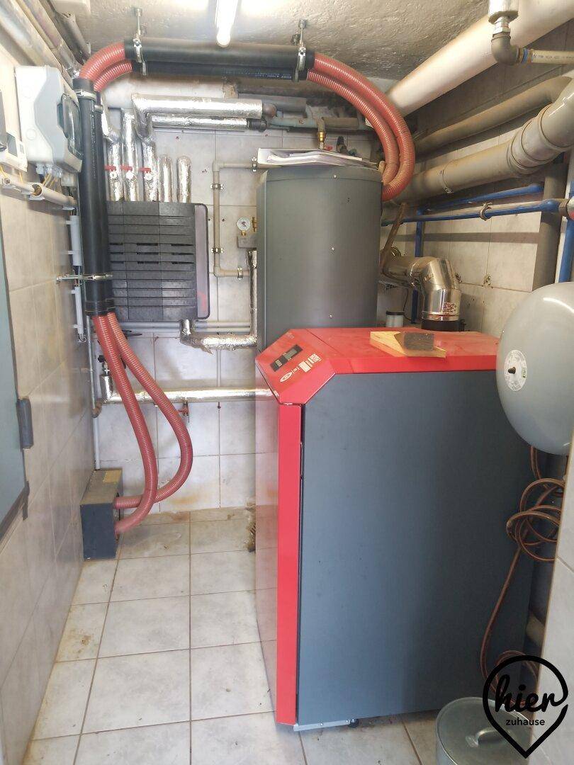Heizungsraum mit großem Heizkessel, Rohren und Warmwasserspeicher auf Fliesenboden.