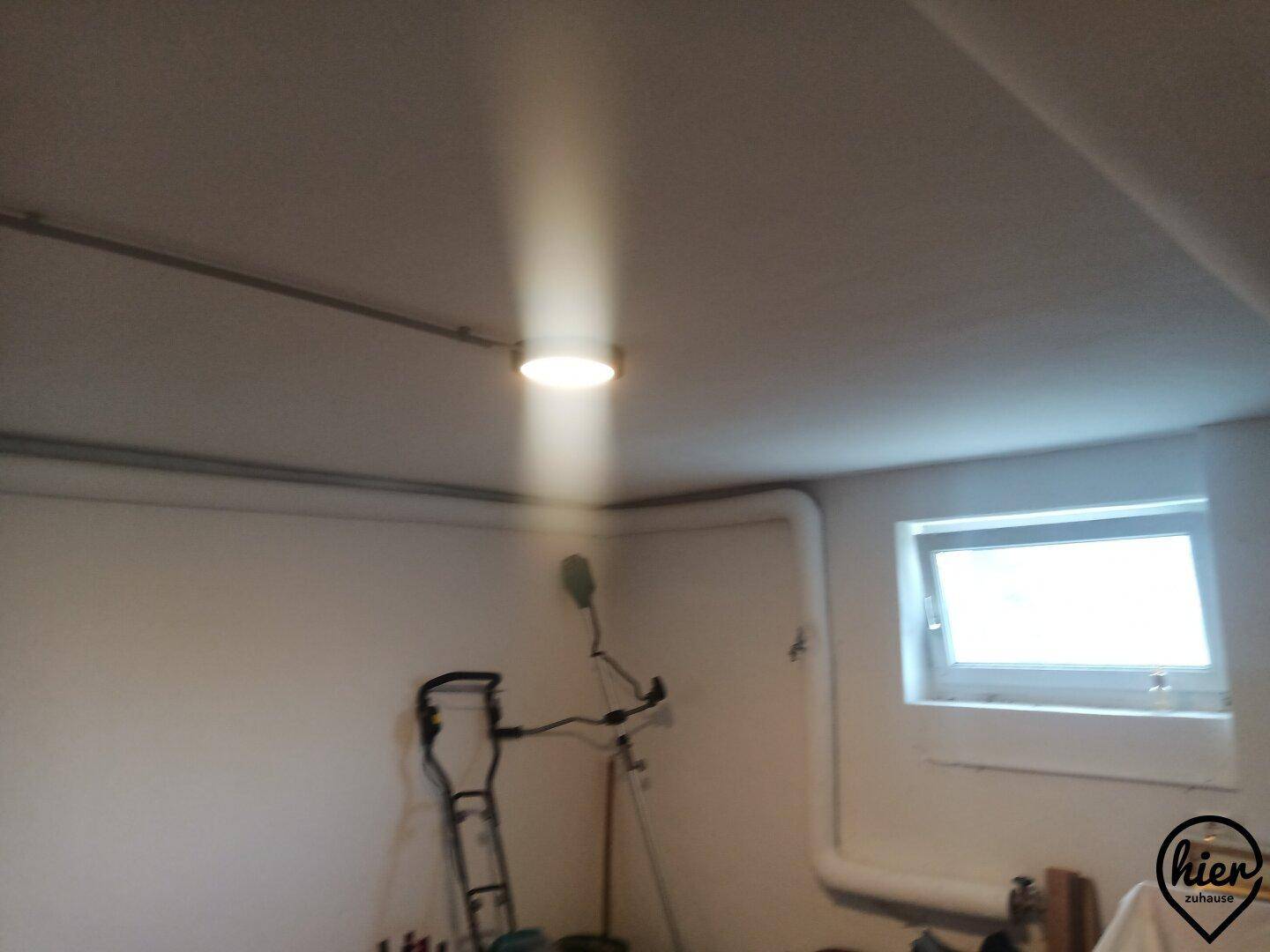 Einfacher Kellerraum mit kleinem Fenster, Deckenleuchte und Stauraum für verschiedene Geräte.