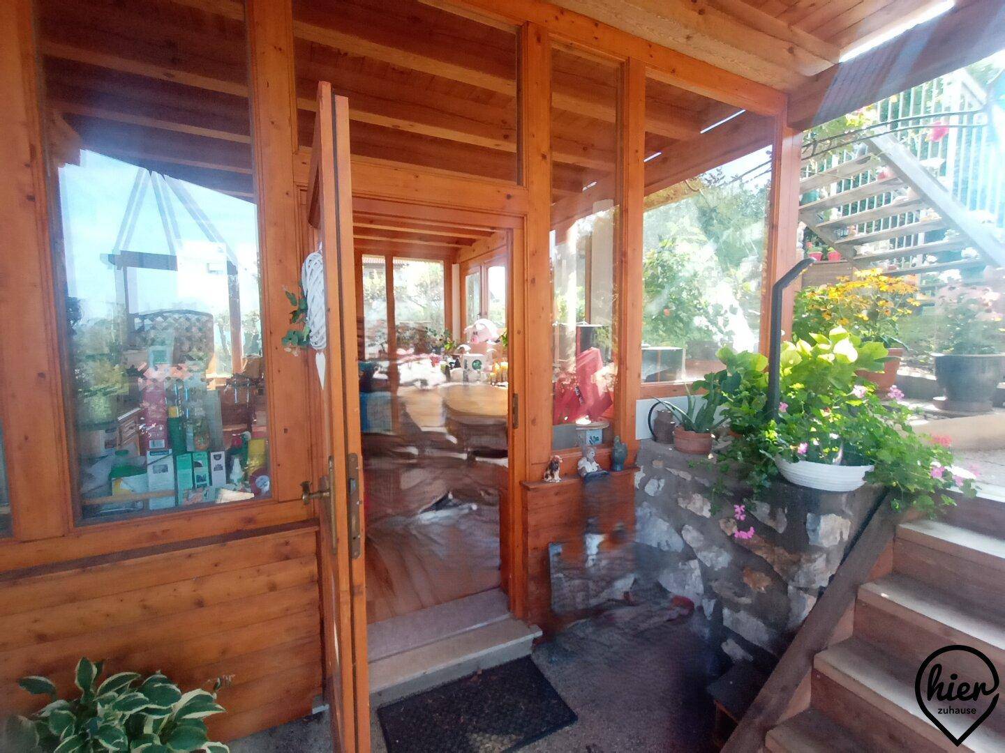 Eingangsbereich zu einem hellen Wintergarten mit Holzverkleidung und Pflanzen im Außenbereich.