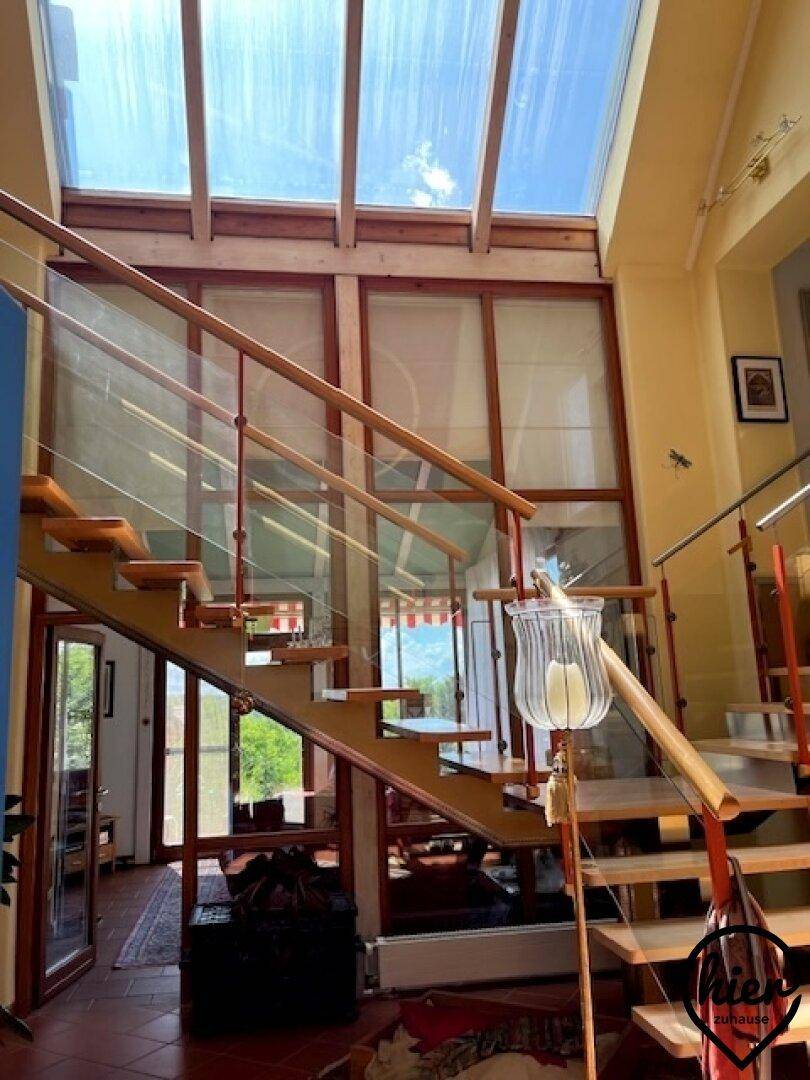 Heller Eingangsbereich mit offener Holztreppe, Glasgeländer und großem Oberlicht für viel Tageslicht.