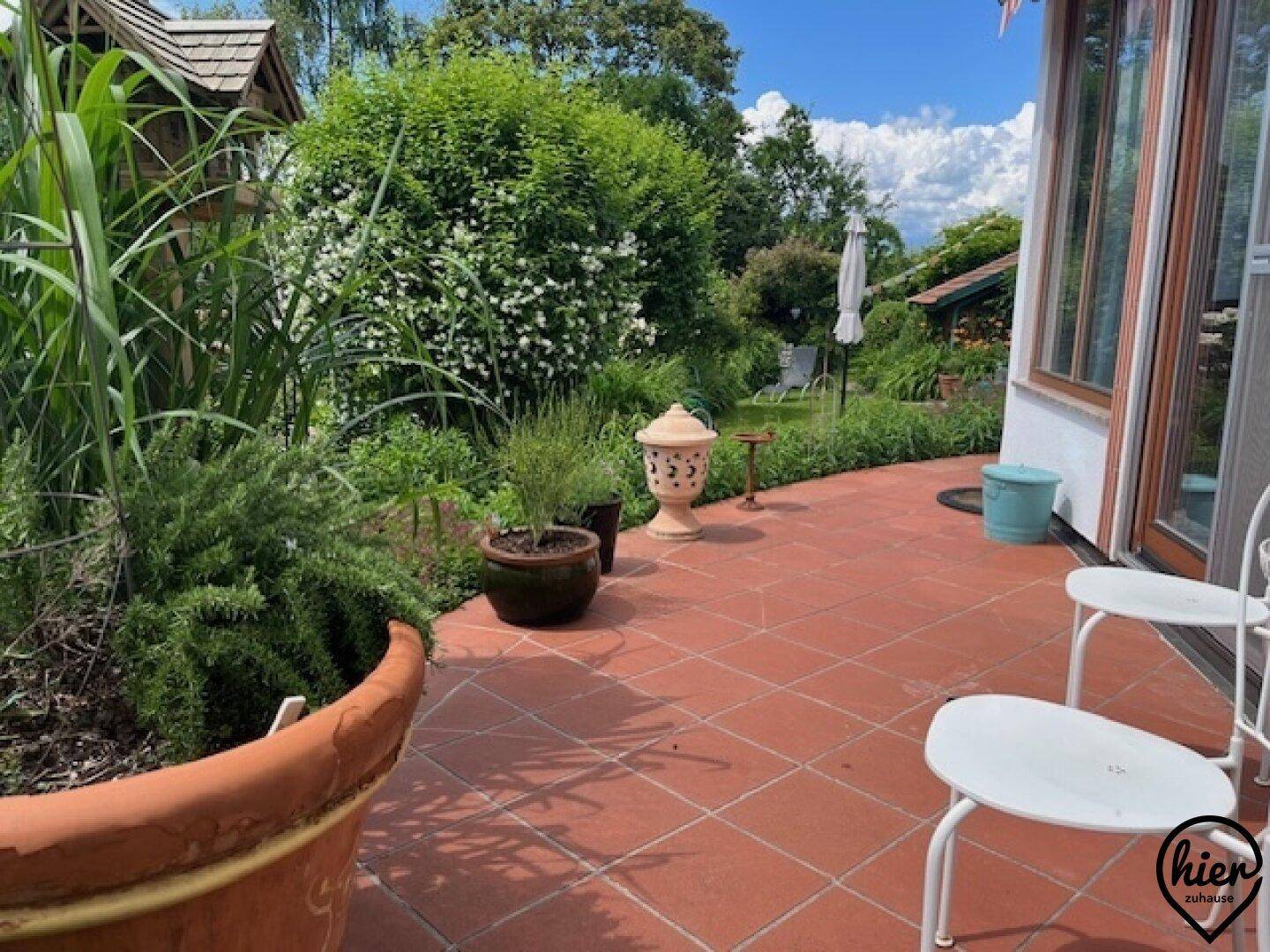 Geräumige geflieste Terrasse mit Gartenmöbeln, Pflanzen und Blick auf den grünen Garten.