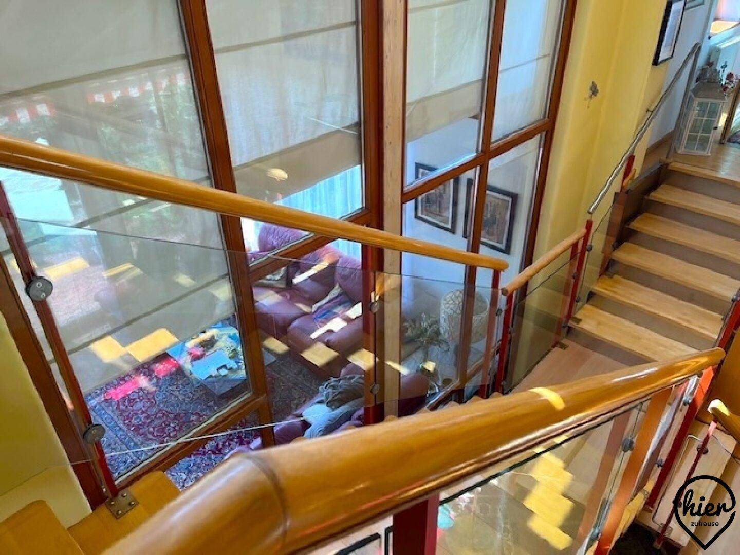 Blick von der oberen Treppe auf das offene Wohnzimmer durch große Fenster und Glasgeländer.
