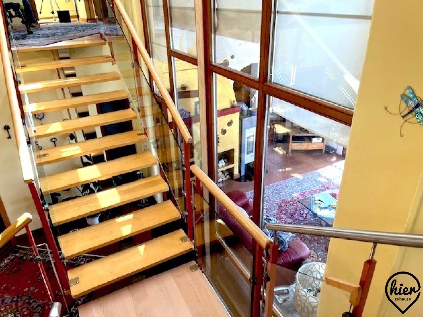 Moderne Holztreppe mit Glasgeländer und Blick in den Wohnbereich durch große Fenster.