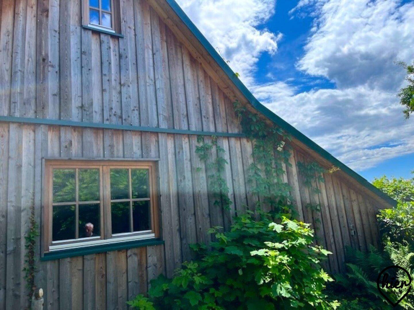 Rustikale Holzfassade eines Hauses mit schrägem Dach und Fenster, umgeben von viel Grün.
