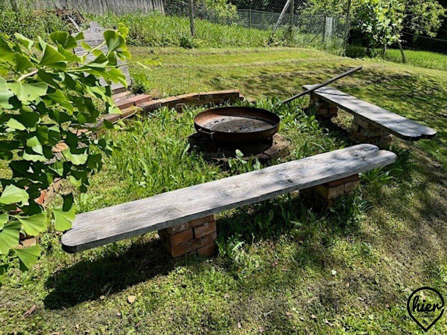 Gemütlicher Sitzbereich im Garten mit Holzbänken und einer Feuerstelle auf der Rasenfläche.