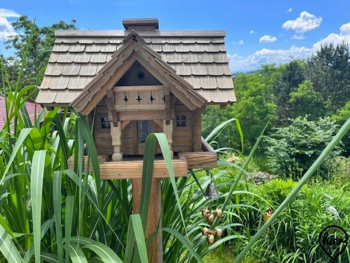 Dekoratives Vogelhaus aus Holz auf einem Pfahl, umgeben von hohem Gras und Grün.