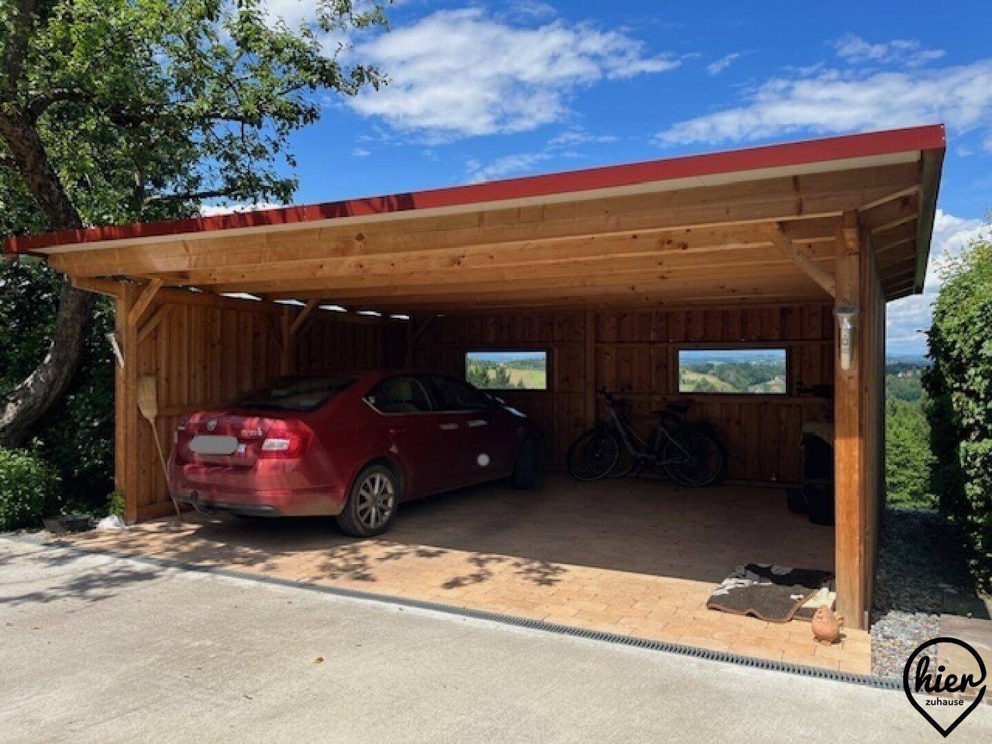 Geräumiger Holzcarport mit rotem Auto und Fahrrädern, umgeben von grüner Natur.