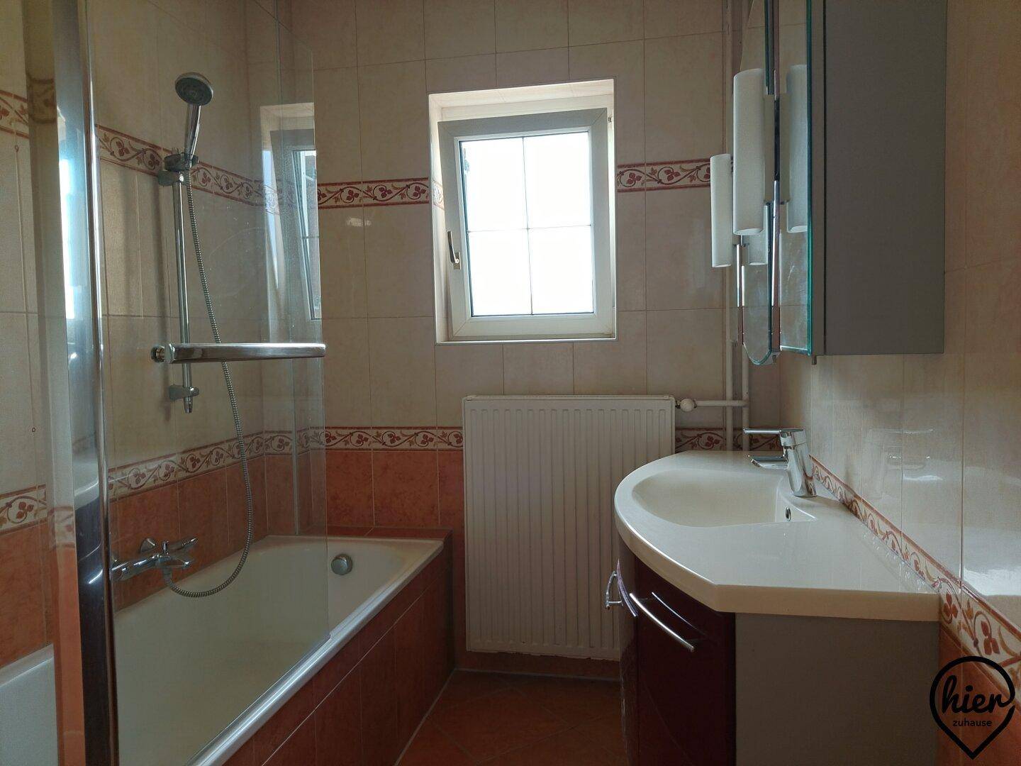 Badezimmer mit Badewanne, Duschabtrennung und Fenster, gefliest in warmen Farbtönen.