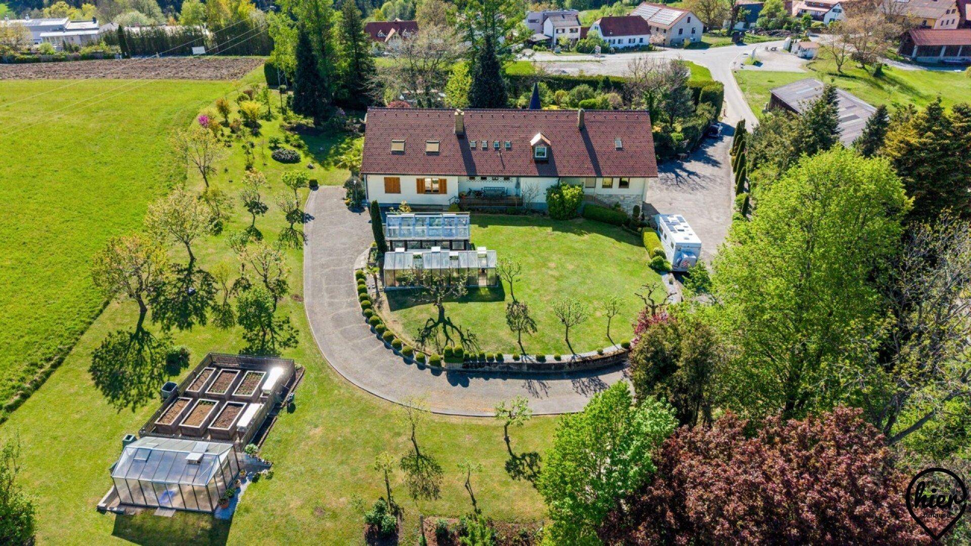 Weitläufiger Gartenbereich mit gepflegtem Rasen, Zierbäumen und mehreren Gewächshäusern, umgeben von Natur.
