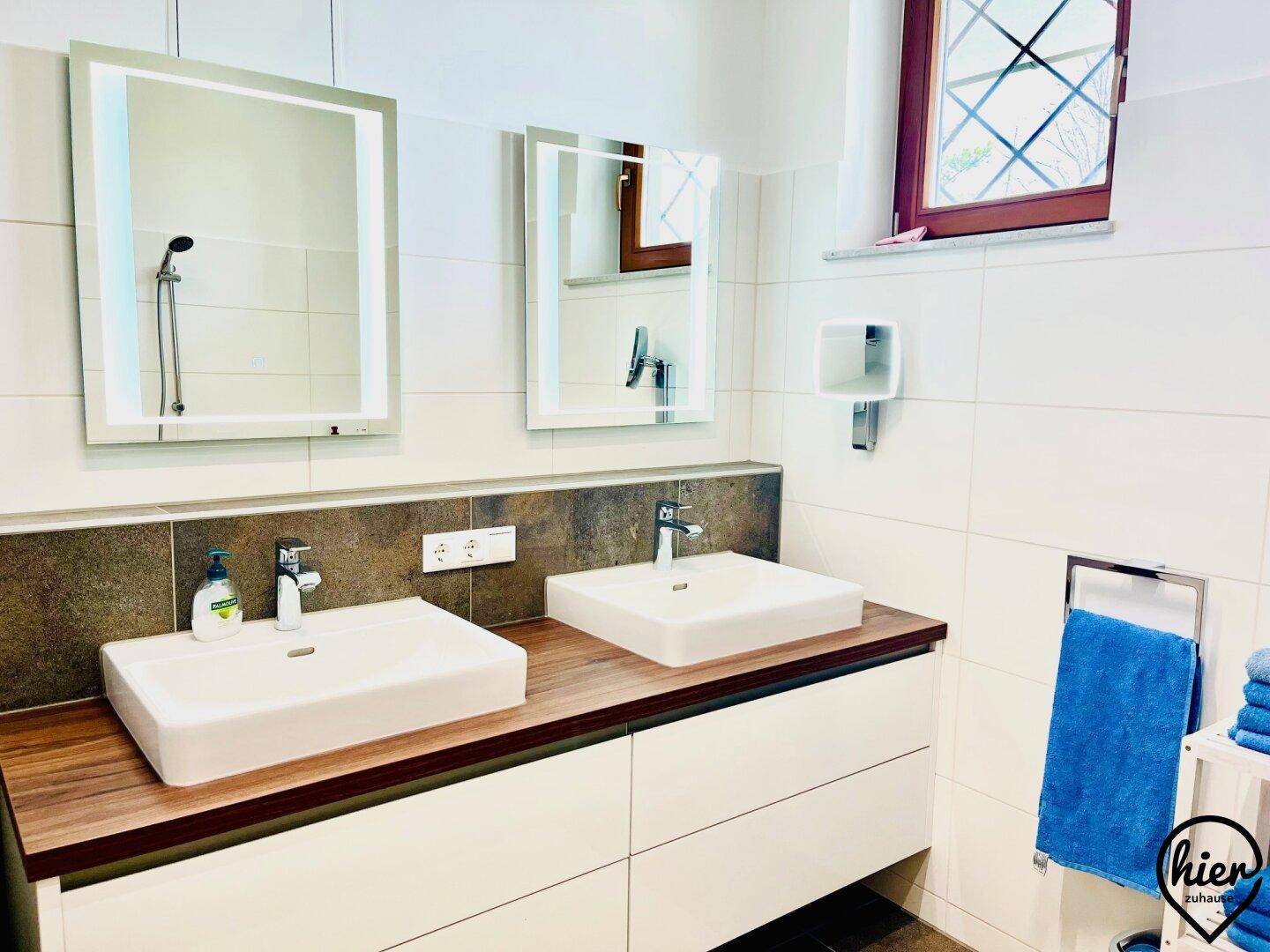 Modernes Badezimmer mit Doppelwaschbecken, beleuchteten Spiegeln und großen weißen Fliesen, sehr gepflegt.
