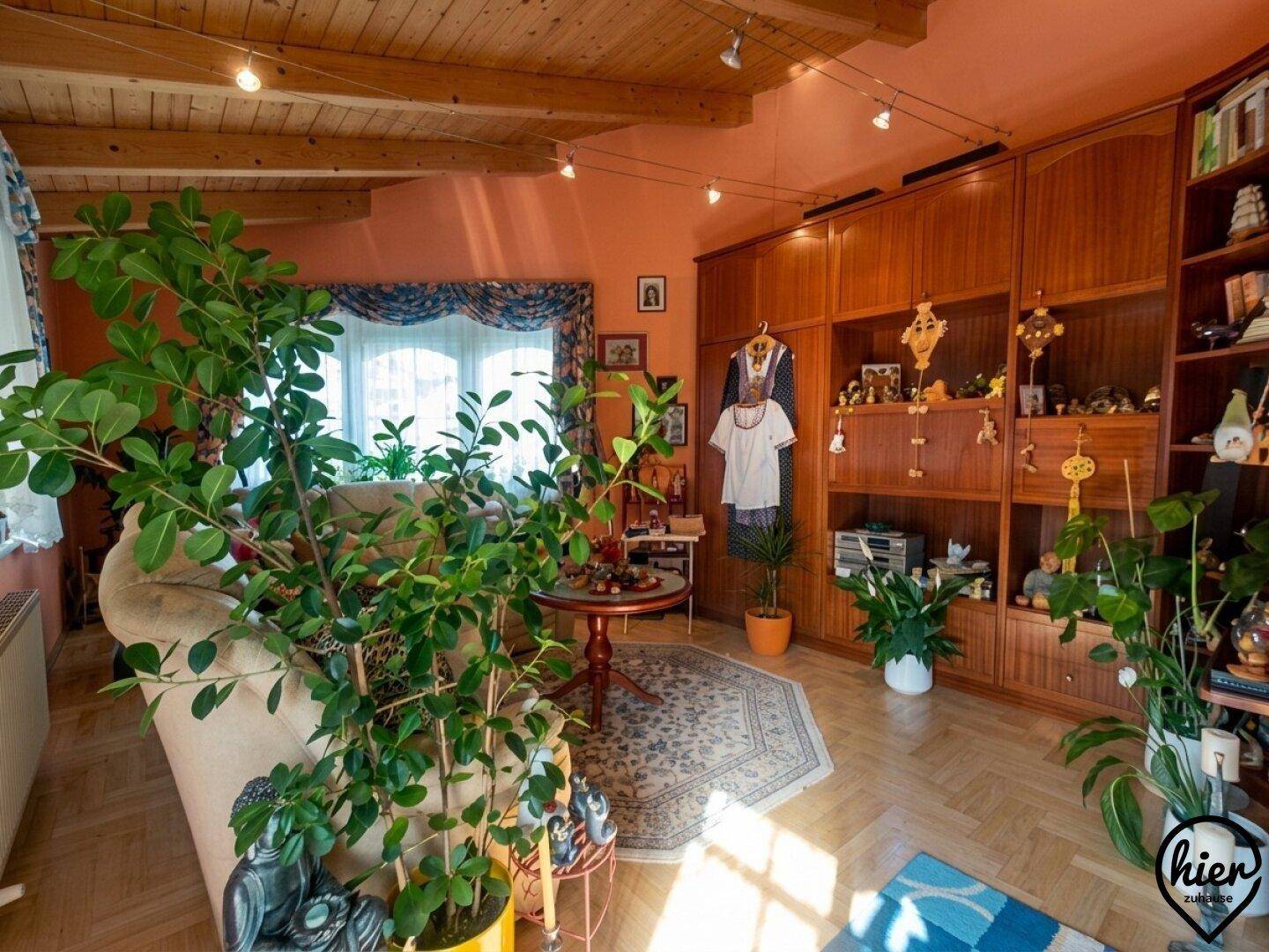 Wohnzimmer mit Holzbalkendecke, vielen Pflanzen und traditionellen Holzmöbeln in warmen Farben.