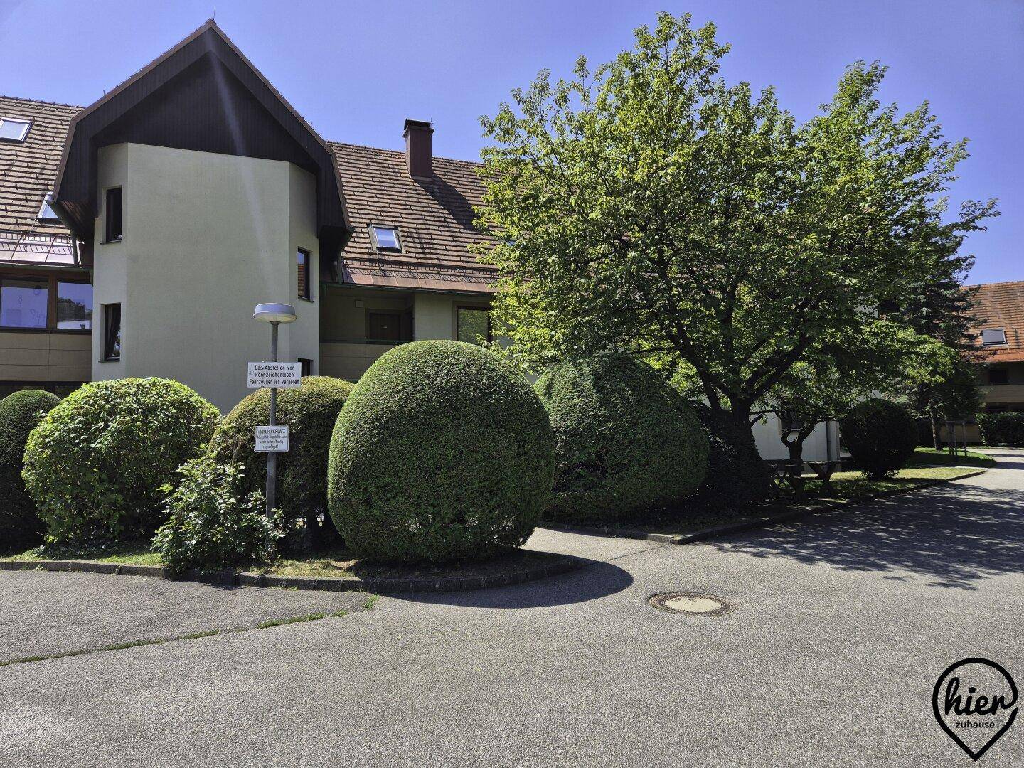 Außenansicht des Gebäudes mit gepflegten Büschen und einem Baum auf dem Vorplatz.