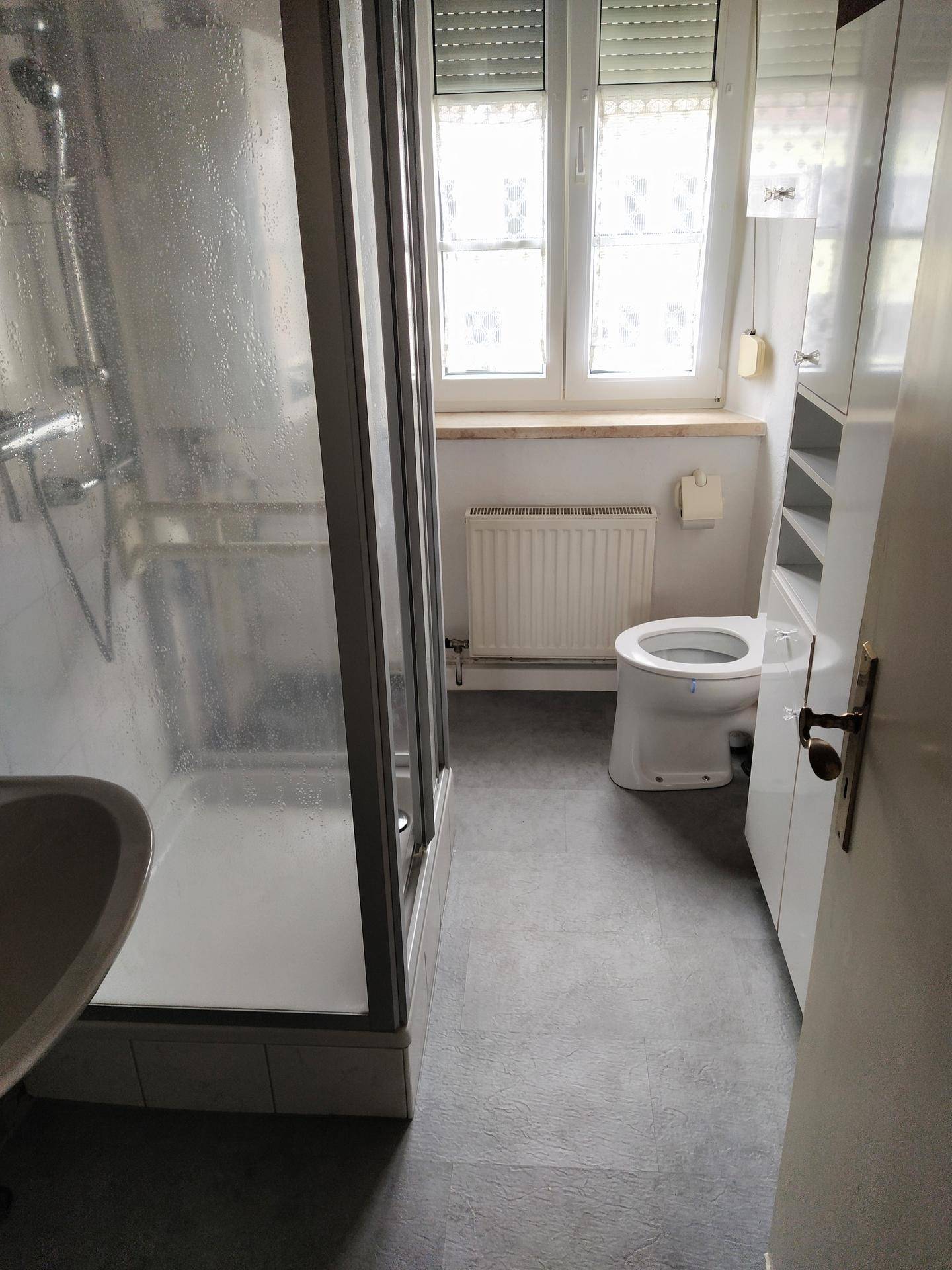 Badezimmer mit Duschkabine, WC, Fenster und grauen Fliesen, in einem gepflegten Zustand.
