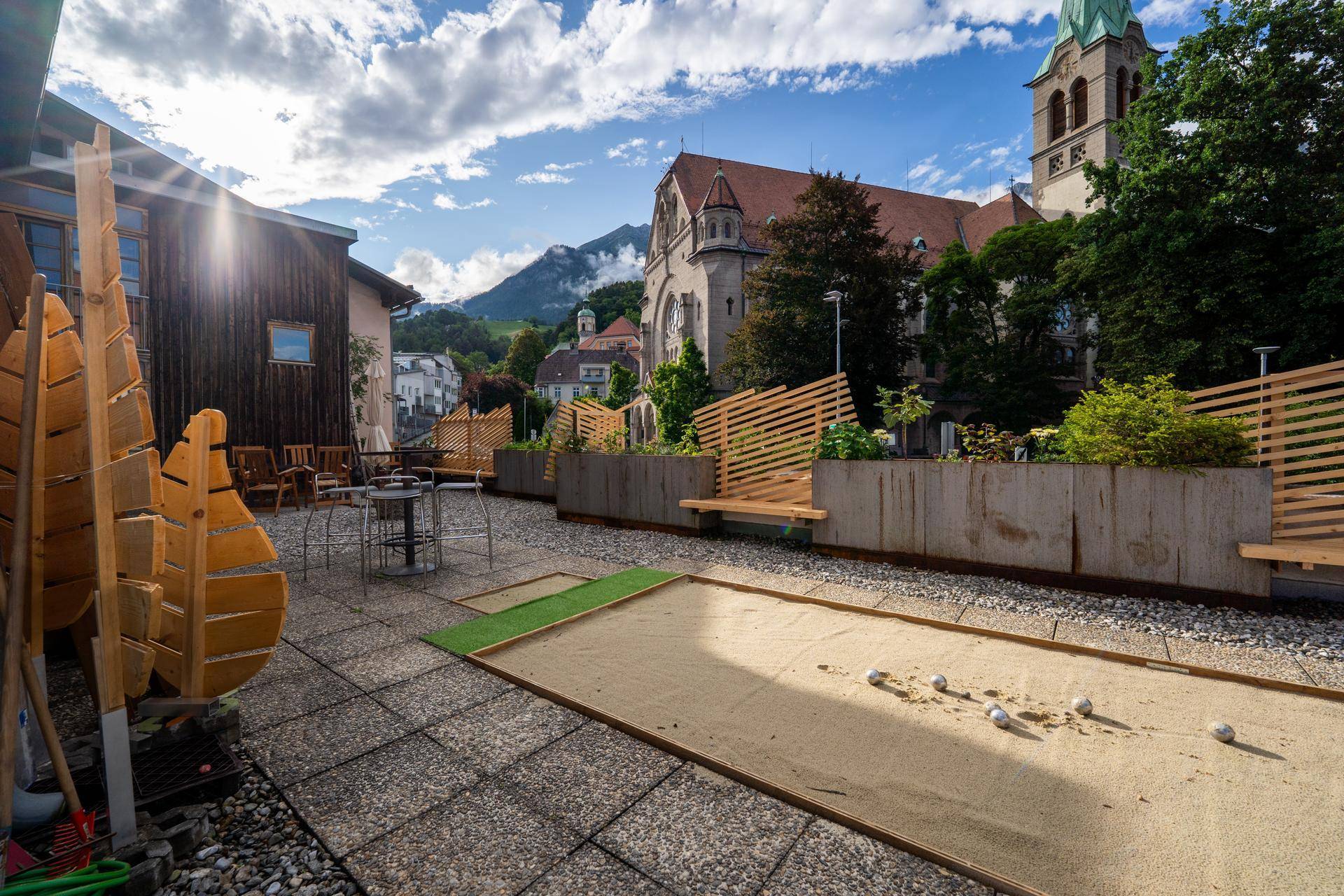 Geräumige Dachterrasse mit Boccia-Bahn, Sitzgelegenheiten und Blick auf eine Kirche und die Berge.