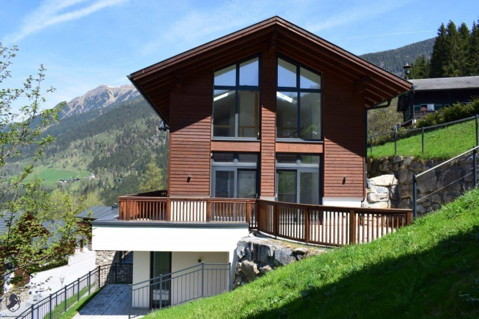 Attraktives Holzhaus mit großen Fenstern und Bergblick in alpiner Landschaft unter blauem Himmel.