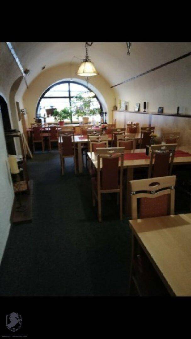 Speisesaal mit gewölbter Decke, Holztischen und einem großen Rundbogenfenster, das Tageslicht hereinlässt.