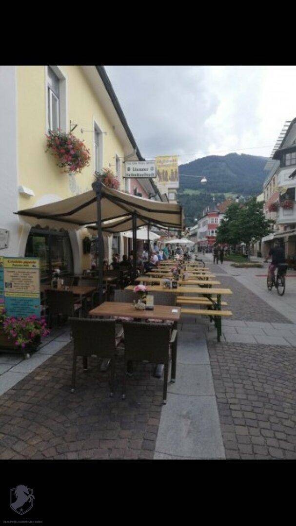 Belebte Fußgängerzone mit Außengastronomie, umgeben von traditionellen Gebäuden und Bergblick im Hintergrund.