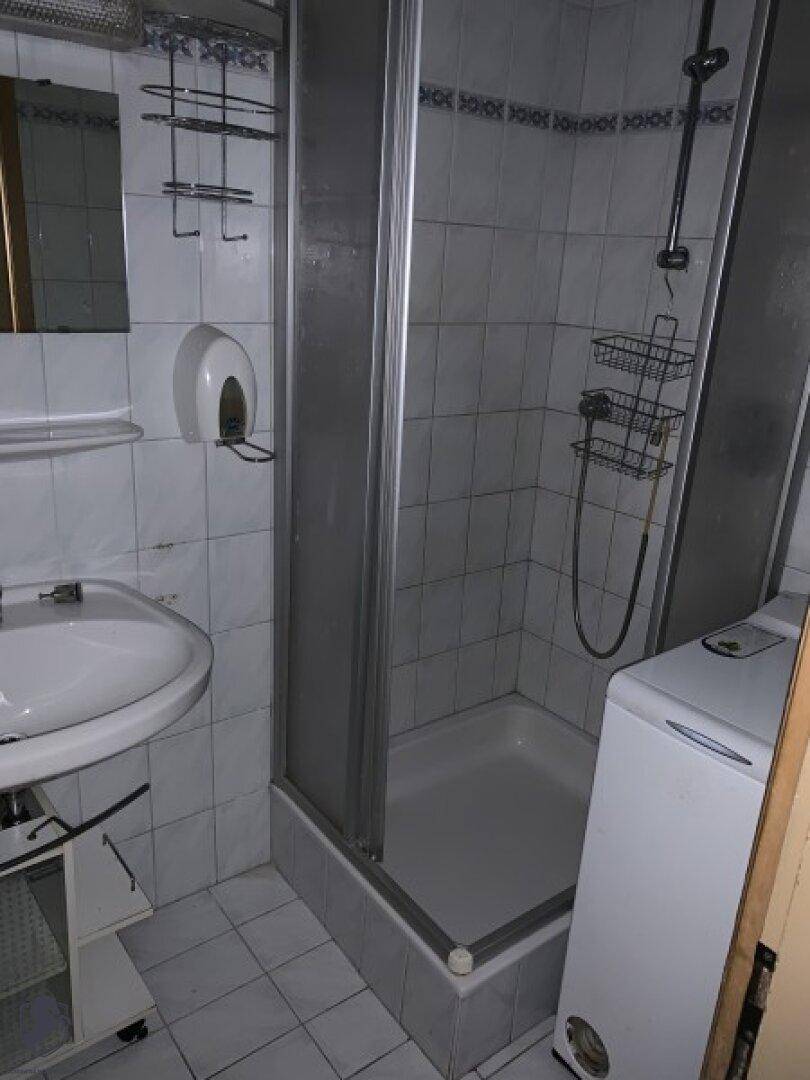 Kleines Badezimmer mit Duschkabine, Waschbecken und weißen Fliesen an Wänden und Boden, inklusive Waschmaschine.