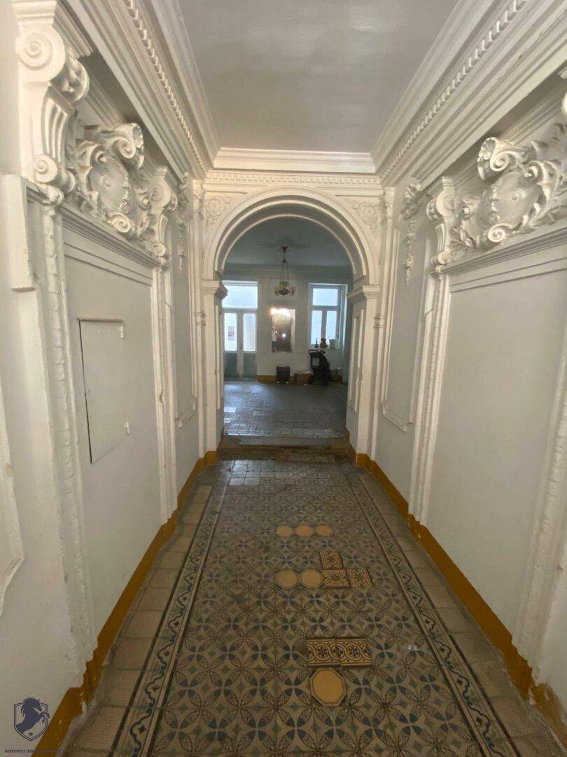 Historischer Eingangsbereich mit aufwendigen Stuckverzierungen und gemustertem Fliesenboden, der in einen weiteren Raum führt.