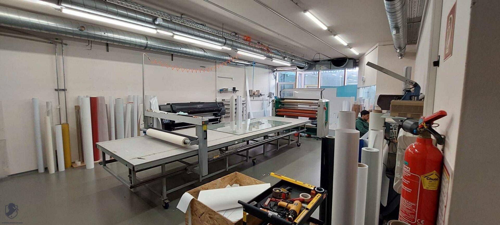Innenansicht einer Werkstatt oder Produktionshalle mit Druckmaschinen, Materialrollen und Arbeitstischen.