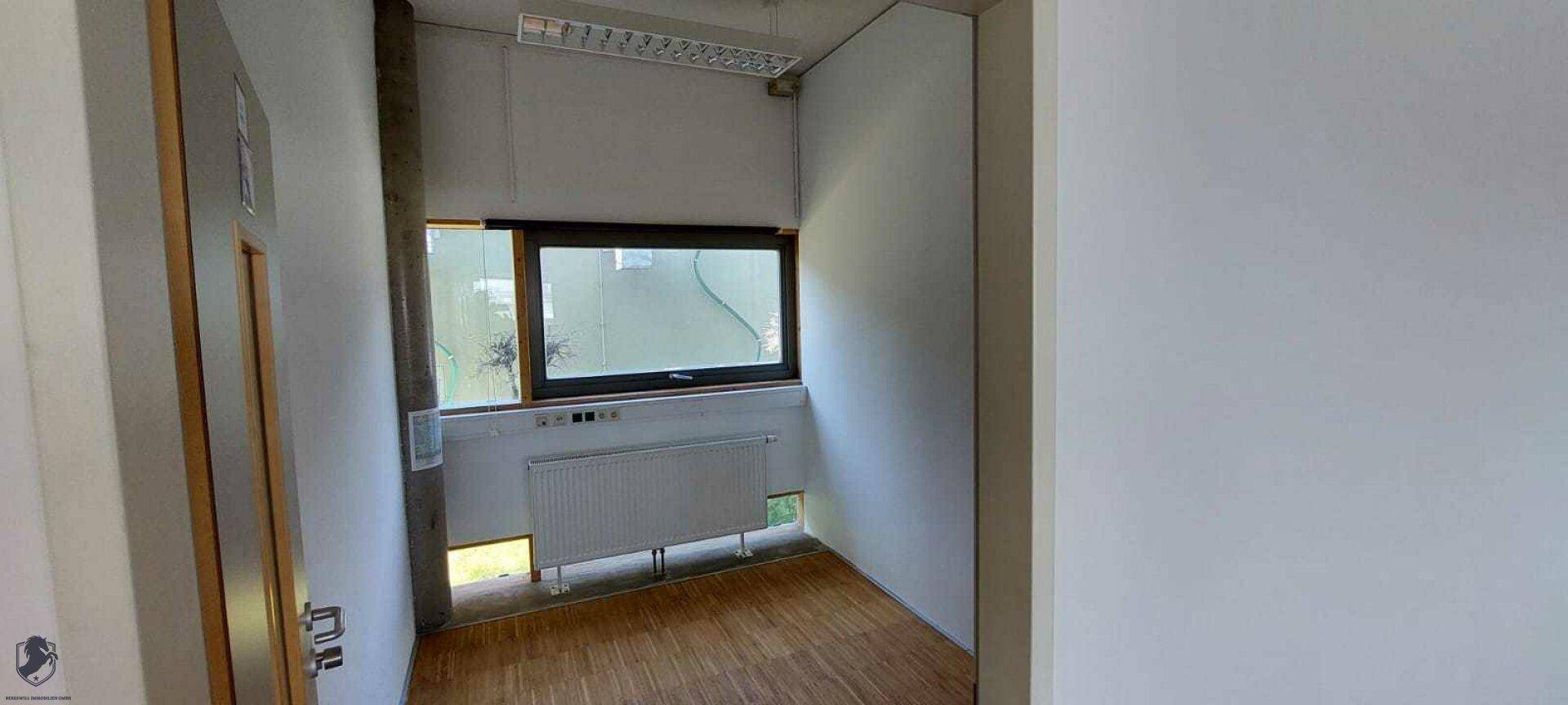 Kleiner, leerer Büroraum mit Fenster, Heizkörper und Holzboden, der viel natürliches Licht erhält.