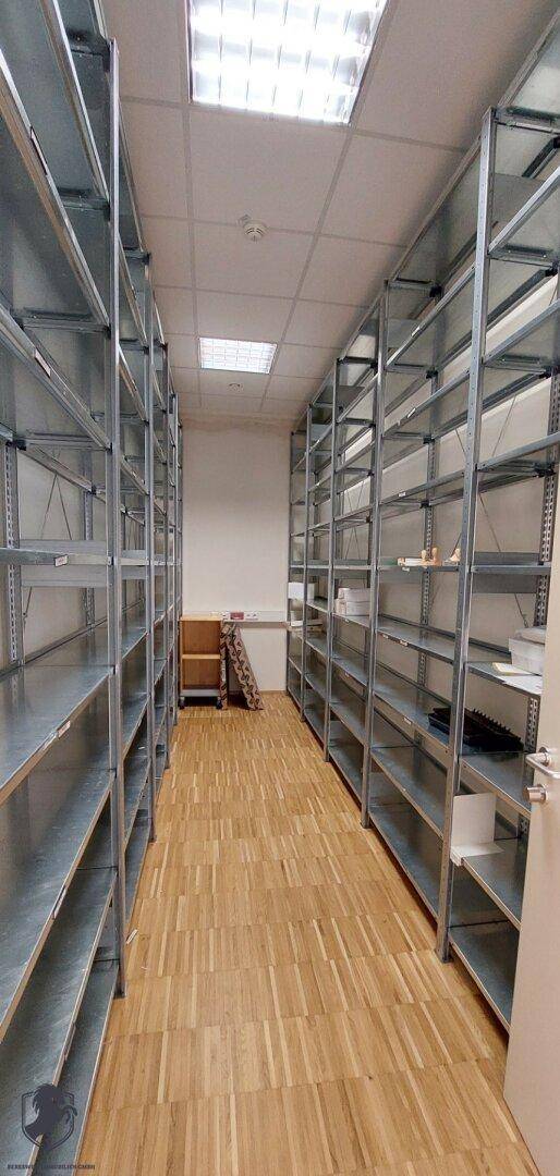 Schmaler Gang in einem Lagerraum, gesäumt von Metallregalen und Holzboden für effiziente Lagerung.