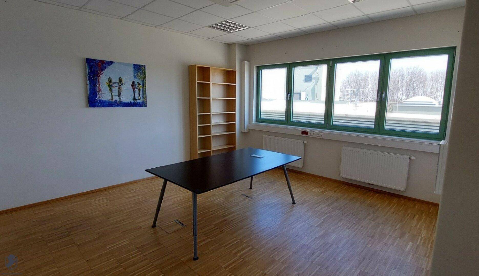 Geräumiger Büroraum mit großem Fenster, Heizkörper, Holzboden und einem modernen Tisch.