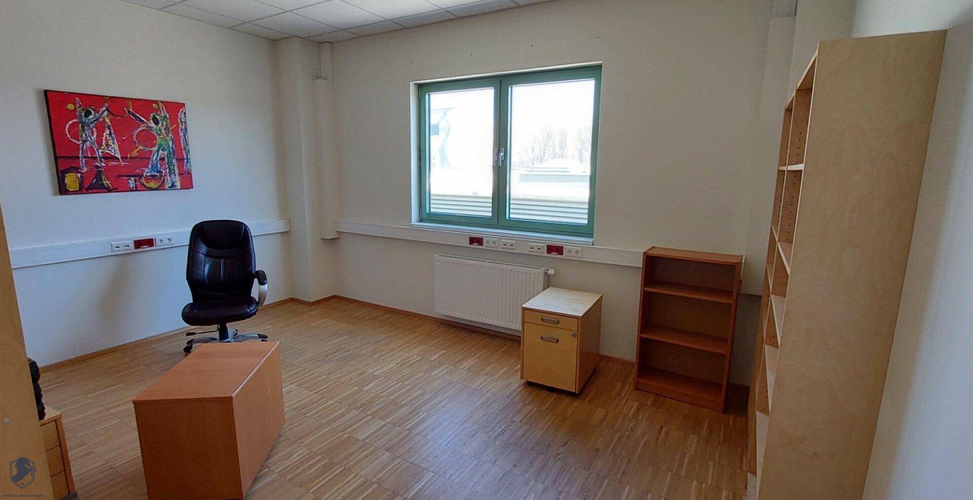 Leerer Büroraum mit Fenster, Heizkörper, Holzboden und einfacher Möblierung, bereit zur Nutzung.