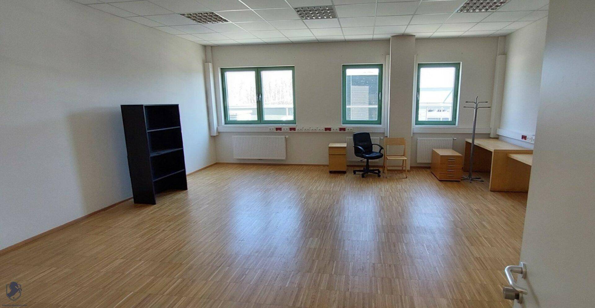 Heller Büroraum mit mehreren Fenstern, Heizkörpern, Holzboden und einfacher Büroeinrichtung.