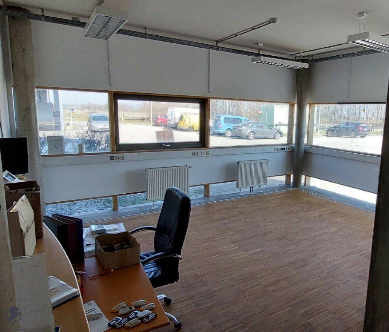 Büroraum mit Schreibtisch, Stuhl und großen Fenstern mit Blick auf den Parkplatz und Fahrzeuge.