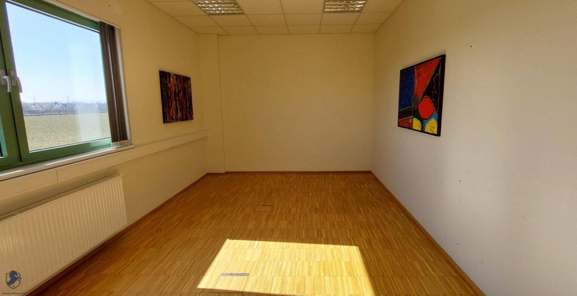 Leerer Raum mit Fenster, Heizkörper, Holzboden und farbenfrohen Kunstwerken an den Wänden.