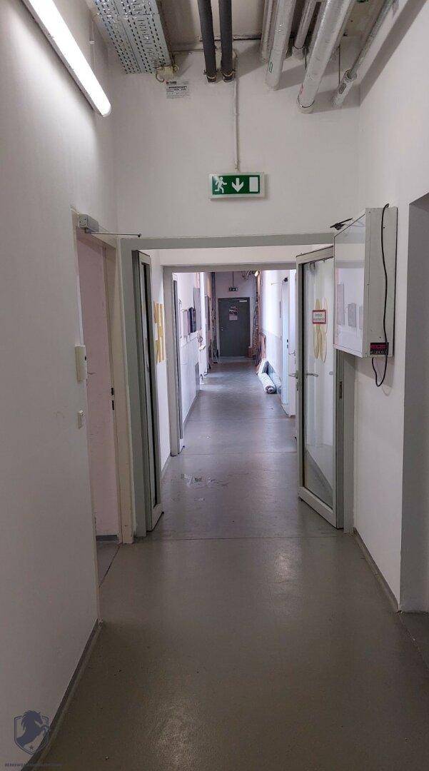 Langer Flur mit grauem Boden, sichtbaren Deckenrohren und einem Notausgangsschild.