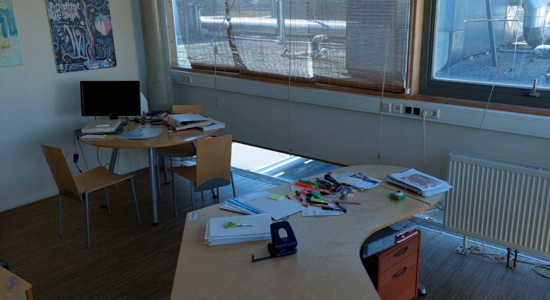 Büroraum mit zwei Schreibtischen, Stühlen und großen Fenstern mit Blick nach draußen.