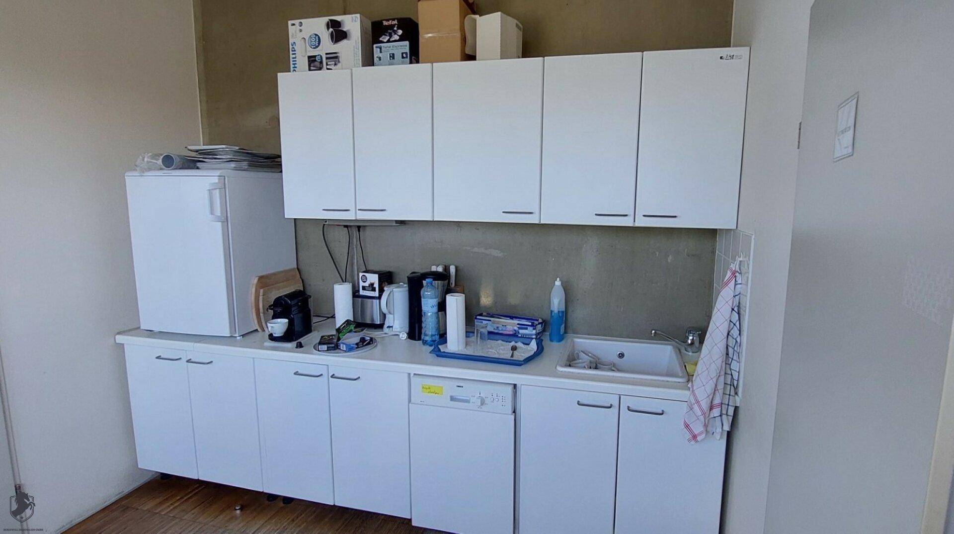 Kleine Küchenzeile mit weißen Schränken, Kühlschrank und Spüle, ideal für Pausenräume.