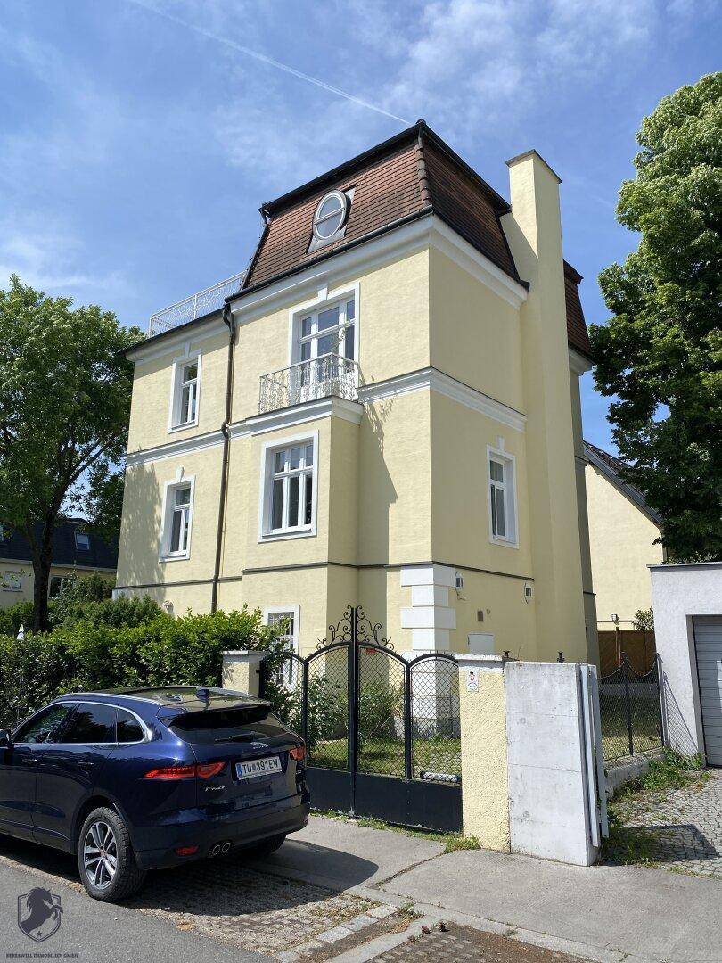 Seitenansicht der gelben Villa mit Mansarddach, Balkon und schmiedeeisernem Tor.